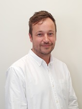 A portrait of James Grayson wearing a business shirt smiling against a plain office wall. 